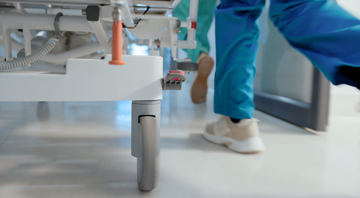 Close-up, low-angle shot of a hospital bed wheel in a hallway with a healthcare worker in blue scrubs walking past, out of focus.